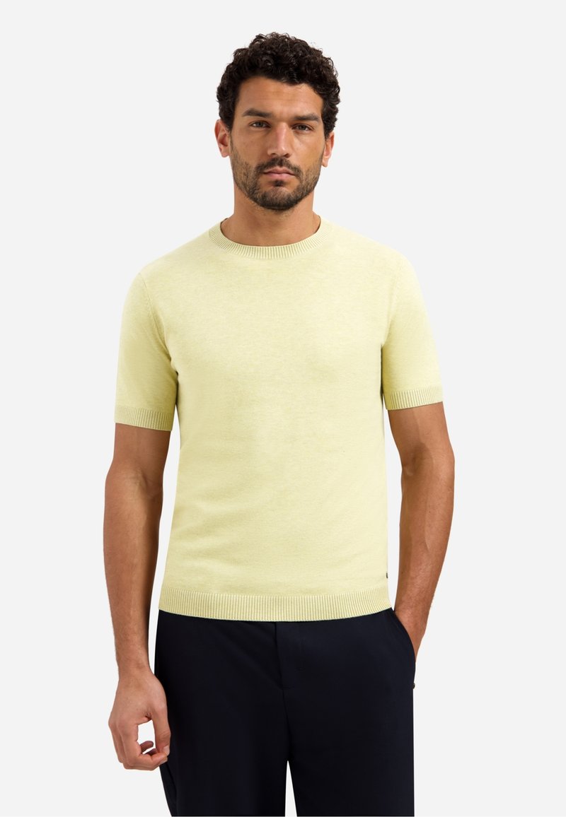 Man met donker krullend haar en baard, gekleed in een lichtgeel kortärmelig truitje en donkere broek, staand tegen een effen achtergrond.