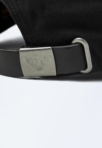 Casquette en coton noir avec une lanière en cuir. La boucle en métal présente des ailes gravées et un logo. Coutures visibles le long des coutures.