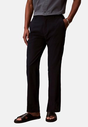 Uomo che indossa pantaloni neri dritti, camicia grigia a maniche corte con bottoni, sandali neri con cinturini incrociati e braccialetto d'argento, mano in tasca.