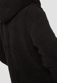 Giacca in pile nera con un esterno testurizzato e morbido. Presenta un cappuccio e una vestibilità rilassata con cuciture visibili lungo le maniche.