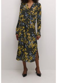 Robe à imprimé floral avec des manches longues, fond vert, fleurs jaunes et bleues, taille nouée et fentes latérales. Complétée par des talons noirs.