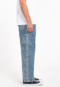 Jeans de mezclilla en un tono azul claro con un corte relajado, que cuentan con un bolsillo lateral, costuras visibles y un leve desgaste. Combinados con zapatillas negras.