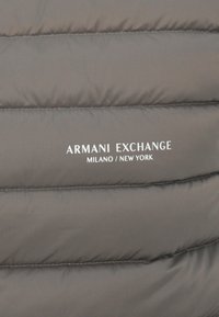 Grå polstret stoff, med horisontale quiltede seksjoner. Merkenavnet "ARMANI EXCHANGE" og "MILANO / NEW YORK" er trykt i hvitt.
