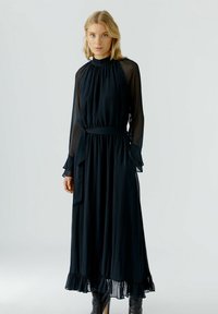 Schwarzes, langärmeliges Kleid aus leichtem Stoff. Mit hohem Kragen, gerafftem Oberteil und gerafftem Saum, der mit einem passenden Gürtel betont wird.