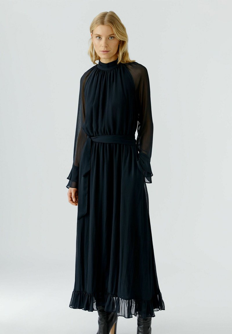 Schwarzes, langärmeliges Kleid aus leichtem Stoff. Mit hohem Kragen, gerafftem Oberteil und gerafftem Saum, der mit einem passenden Gürtel betont wird.