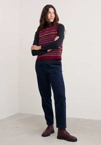 Raudonas mezginys liemenė su daugiaspalve geometrine rašykla, uždėta ant juodos ilgomis rankovėmis marškinėlių. Jūrinės velveto kelnės ir vyšniniai batai.