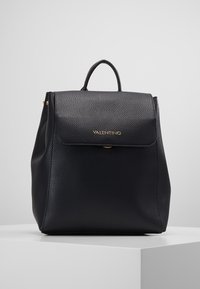 Mochila de cuero negro con una superficie texturizada, solapa plegable y acentos de hardware dorado. Presenta un asa superior y una forma redondeada.