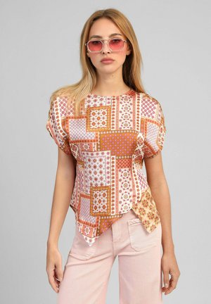 Jeune femme portant un haut à manches courtes à motifs géométriques orange et rose et un pantalon rose clair, avec des lunettes de soleil teintées roses.