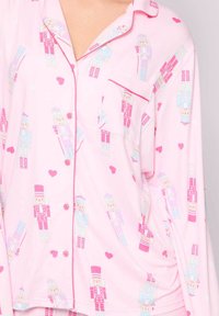 Camisa de pijama rosa con patrón de cascanueces. Cuenta con cierre de botones, bolsillo y un ribete rosa en contraste en el cuello y los puños. Textura de tela suave.