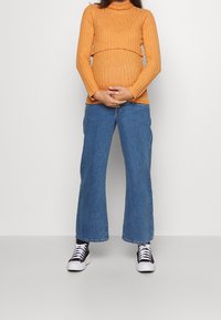 Oranžový žebrovaný rolák, světle modré široké džíny a černobílé tenisky. Ruce spočívající na zaobleném břiše.