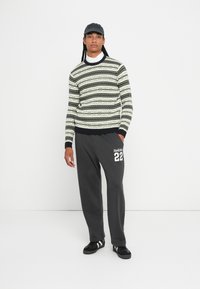 Man met lange vlechten die een gebreide trui met patroon draagt, een witte overhemd met kraag, donkere trainingsbroek met "Hollister 22," zwarte sneakers en een zwarte pet.