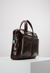 Borsa da lavoro in pelle marrone con due tasche frontali con zip, accentata da dettagli blu e dotata di manici doppi. Texture liscia e design strutturato.