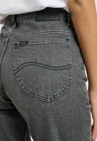 Vaqueros de mezclilla gris con un diseño estilizado en el bolsillo trasero, etiqueta de cuero en la cintura y detalles visibles de costura.