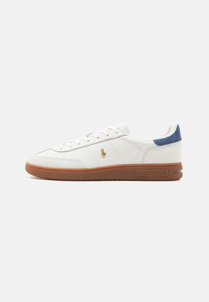 Zapatilla blanca de caña baja con suela marrón, logotipo dorado de Polo Ralph Lauren en el lateral, lengüeta de talón azul y cordones blancos sobre un fondo liso.
