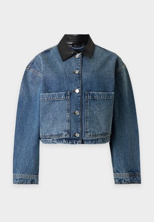 Jeune femme debout portant une veste en jean foncée avec col noir, un jean large assorti, un haut blanc et des chaussures à talons noires sur un fond uni.