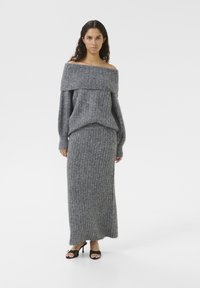 Grå stickad off-shoulder tröja i kombination med en matchande lång ribbad kjol, med mjuk textur och avslappnad passform. Bärs med svarta klackskor.