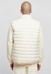 Gepolsterte Weste in Off-White, mit hohem Kragen und horizontaler Steppmusterung; getragen über einem passenden hellfarbenen Sweatshirt.