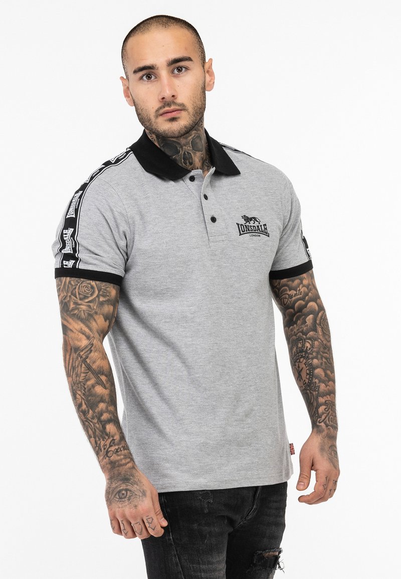 Hallide gris polo särk musta kraega, millel on logo ja mustrilised aktsendid varrukatel. Mudel kannab musti teksaseid, näidates tätoveeringuid oma kätele.