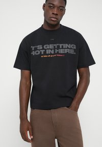 Νέος άνδρας με μαύρη μπλούζα T-shirt που έχει γκρίζο και κόκκινο κείμενο "ΓΙΝΕΤΑΙ ΖΕΣΤΑ ΕΔΩ" και καφέ παντελόνι, που στέκεται μπροστά σε απλό λευκό φόντο.