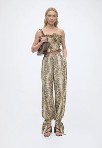 Γυναίκα που φοράει μπεζ κορσέ με φλούδες φιδιού και βολάν και παντελόνι σετ, κρατώντας καφέ τσάντα ώμου, στέκεται μπροστά σε λευκό φόντο.