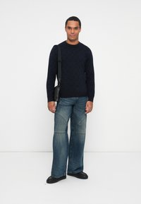 Hombre vistiendo un suéter de patrón oscuro, jeans de pierna ancha azules, zapatos negros y llevando un bolso negro de hombro frente a un fondo blanco liso.