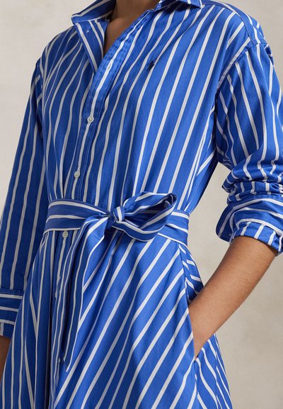 Inimene, kes kannab sinist ja valget vertikaalsete triipudega nööbitavat kleiti vööga, mille varrukad on üles keeratud, ja käsi taskus.