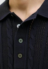 Tricou polo bleumarin cu un model de tricot texturat și un design cu cabluri, două butoane verzi și o guler țesut. Vizualizare de aproape.