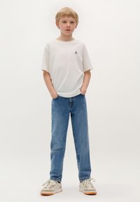 T-shirt bianca in cotone con una piccola grafica scura, abbinata a jeans in denim azzurro chiaro e sneakers beige con suole marroni. Postura in piedi.