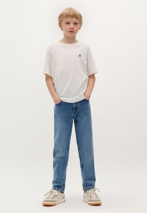 T-shirt bianca in cotone con una piccola grafica scura, abbinata a jeans in denim azzurro chiaro e sneakers beige con suole marroni. Postura in piedi.