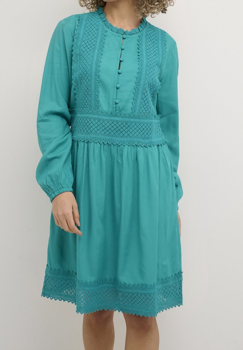 Robe bleu sarcelle à manches longues, devant boutonné, avec des détails en dentelle élaborée sur le corsage et l'ourlet. Tissu doux avec une silhouette ample.
