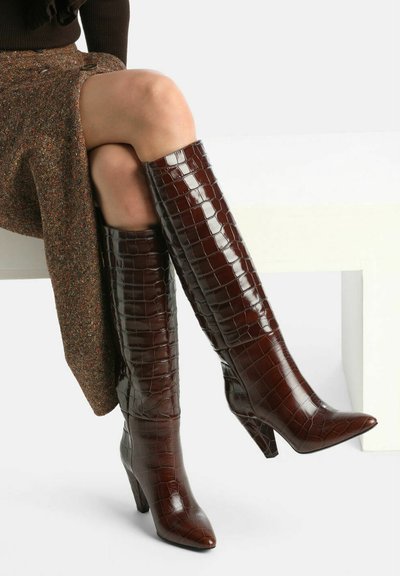 Botas de cuero color marrón con relieve de cocodrilo, hasta la rodilla, con puntas afiladas y tacones altos, que presentan un acabado brillante y una silueta elegante.