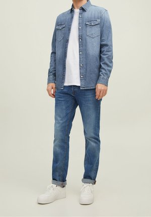 Camisa de mezclilla azul con botones y dos bolsillos en el pecho, llevada sobre una camiseta blanca y combinada con vaqueros azules y zapatillas blancas.
