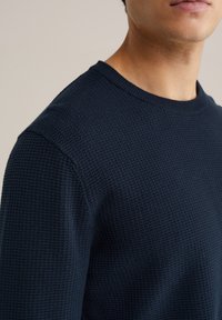 Tengerészkék kötött pulóver texturált anyagból, kerek nyakkivágással és hagyományos fazonnal. Közeli kép a váll és a mellkas területéről.