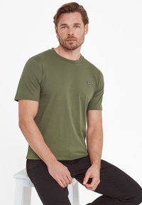 Olive green t-shirt in a soft, cotton material with a round neck, short sleeves, and a small logo on the chest. Worn with dark pants.