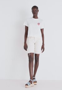 T-shirt branca de manga curta com um gráfico de morango vermelho, combinada com calções de ganga cor creme e sandálias de plataforma azuis. Tecido suave.