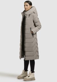 Vrouw in een lange beige gewatteerde jas met capuchon, zwarte leggings en beige plateauboots, staand met de handen in de jaszakken.