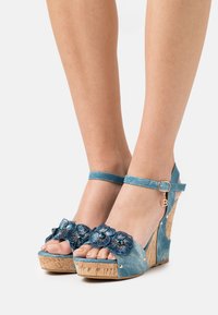 Blå denim kilklackar med korkplattform, blommor som dekoration, ankelsnöre med spänne och liten varumärkescharm.