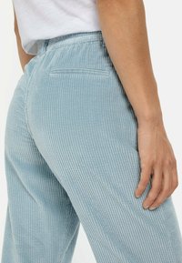 Lysblå corduroy bukser med vertikal ribbing, elastisk talje og en enkelt baglomme. Blød tekstur synlig i stofdetaljer.