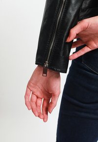 Rukáv černé kožené bundy s rukávem na zip. Zip má kovový jezdec. Ruka umístěná blízko bundy, ukazující tón pokožky.