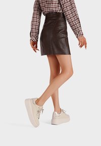 Mini gonna in finta pelle marrone con vita alta e texture liscia, abbinata a sneakers a piattaforma color crema con un accento lucido sul tacco.