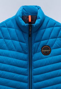 Blå quiltad jacka med hög krage, framsidans dragkedja och ett runt logotypmärke på vänster bröst. Har en slät, glänsande yta.