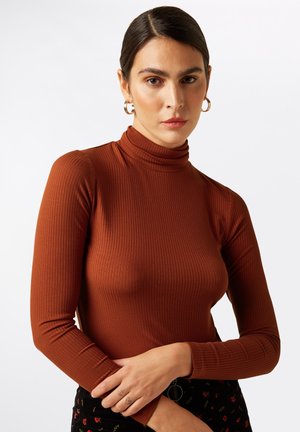 Maglia a maniche lunghe a collo alto a costine in tessuto marrone caldo; presenta un colletto alto e un design aderente; indossata con una gonna nera a fantasia.