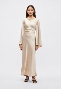 Ilga satino suknelė smėlio spalvos, su aptemptu liemeniu ir laisvai krentančia suknele, šiek tiek blizgi ir lengvai krentanti. Juodi aukštakulniai su dirželiais puikiai papildo įvaizdį.