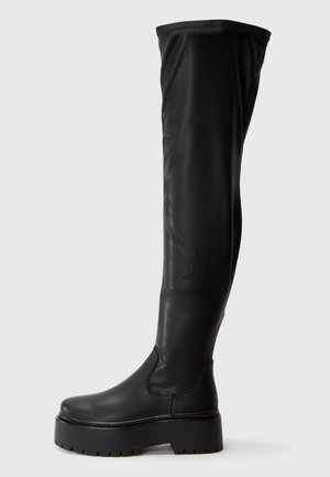 Botte haute noire en cuir avec plateforme, bout arrondi et semelle épaisse sur fond gris clair uni.