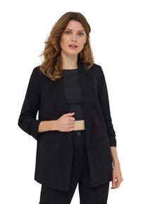 Blazer nero su misura con revers, maniche lunghe, due tasche frontali e una finitura in tessuto liscio. Indossato sopra un crop top nero.
