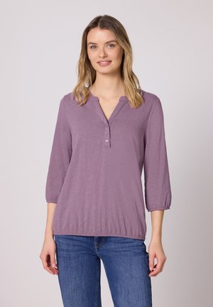 Giovane donna con capelli biondi di lunghezza media, che indossa una camicetta viola con tre bottoni e jeans blu, in piedi davanti a uno sfondo chiaro semplice.