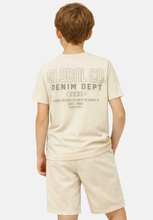 Jongen met kort bruin haar, draagt een beige T-shirt met de tekst "GLOBAL CO. DENIM DEPT" en bijpassende korte broek, staat met zijn rug naar de camera.