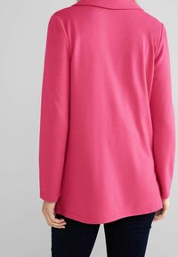 Fuksian va pitkähihaine paita, jossa on sileä rakenne, kaulus ja kaareva helma, yhdistettynä tummiin farkkuihin.