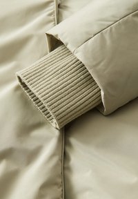 Giacca khaki imbottita con dettagli a maniche a coste; superficie liscia e opaca con una leggera lucentezza sul tessuto esterno.