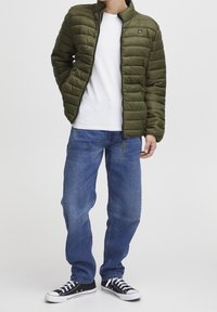 Olivgrüne Steppjacke mit Steppmuster, Reißverschluss vorne, getragen über einem weißen Hemd, kombiniert mit blauen Jeans und schwarzen Sneakern.